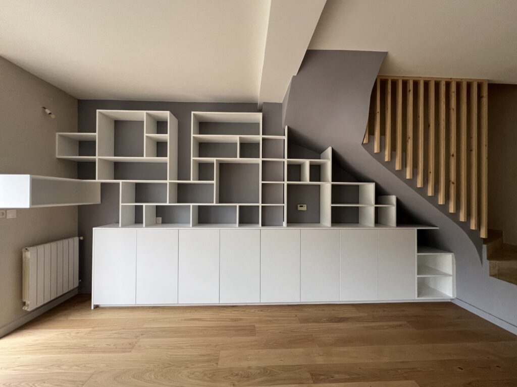 Structure bibliothèque asymétrique sur mesure blanc sur mur gris sous escalier avec claustra garde corps en bois massif.