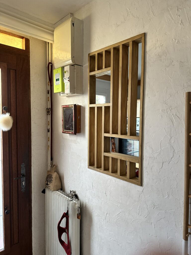Création sur mesure de claustra bois encastré dans un mur d'entrée, offrant une vue sur une cuisine lumineuse.