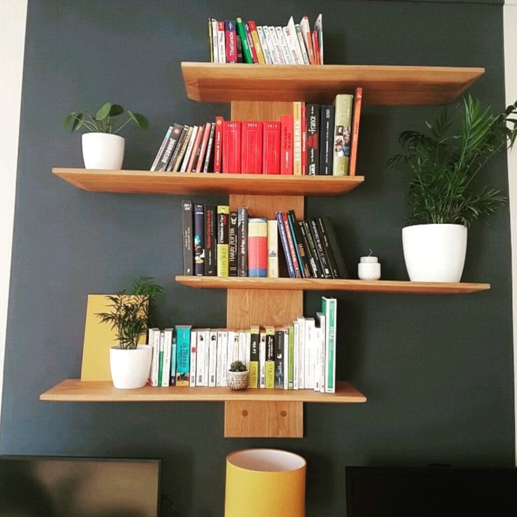 etagere-bois-montant-central Étagère murale asymétrique en bois naturel sur un mur sombre, décorée de livres colorés et de plantes d'intérieur en pots blancs.